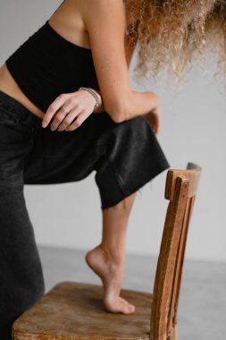 Kıvırcık saçlı, koyu renk bluzlu ve gri arka planda tahta koltuğun yanında poz veren bir kadın.