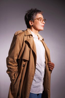 Trençkotlu, gözlüklü ve beyaz gömlekli zarif, olgun bir kadın. Gri üzerinde yalnız poz veriyor.