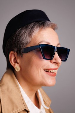 Gri renkli güneş gözlüklü, bereli ve trençkotlu gülümseyen olgun kadın portresi 