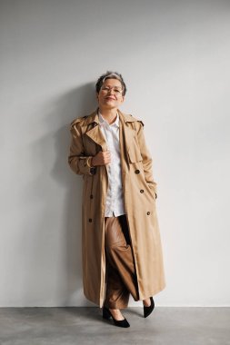 Gri duvarın yanında gözlüklü ve trençkotlu şık ve gülümseyen olgun bir kadın.