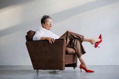 Beyaz tişörtlü, kırmızı topuklu ve gri koltukta oturan zarif ve kendinden emin olgun bir kadın.