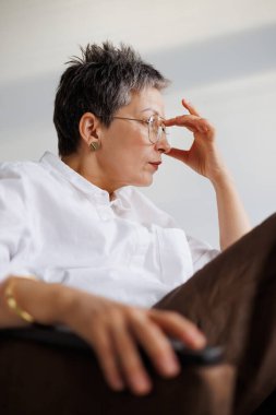 Beyaz gömlekli, gri saçlı bir kadının portresi gri arka plandaki koltukta gözlüğe dokunuyor.