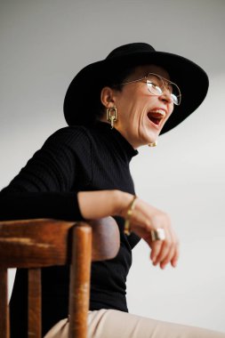 Gözlüklü ve siyah şapkalı heyecanlı ve şık olgun kadın gri sandalyede yalnız başına oturuyor. 