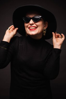 Kırmızı dudaklı, güneş gözlüklü ve siyah şapkalı neşeli, olgun bir kadın. 