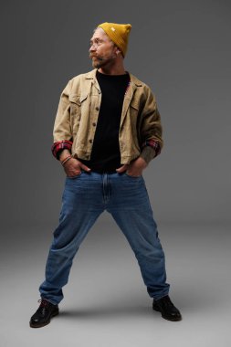 Şapkalı şık sakallı adam, ceketli ve gri üzerinde poz veren kotlu. 