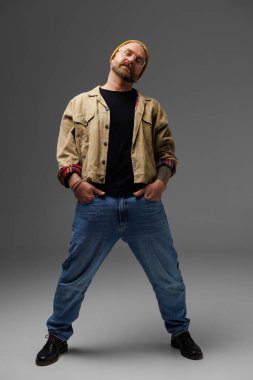 Gözlüklü, kendine güvenen ve şık dövmeli bir adam. Gri ceketli ve kot pantolonlu. 