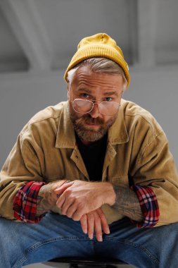 Şapkalı, sıradan ceketli ve stüdyoda kameraya bakan şık dövmeli adamın portresi.