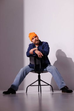 Şapkalı ve rahat gömlekli şık sakallı bir adam. Gri sandalyede oturuyor, gölgesi var.