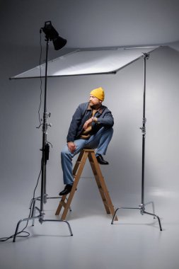 Şapkalı şık sakallı adam gri ışıkta merdiven üstünde poz veriyor.