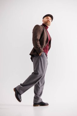 Düz şapkalı, balıkçı yakalı ve ceketli, gri arka planda yürüyen kendine güvenen sakallı adam.