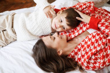 Süveter giyen çocuk yatakta canlı hırka giyen gülümseyen bir anneyle konuşuyor.