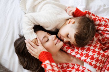 Süveter giyen çocuk annesinin kafasına dokunuyor. Yatakta yatarken, üst tarafta.
