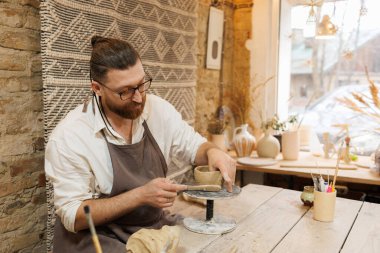 Sakallı bir zanaatkar atölyedeki kil bardağı modelleme aracıyla şekillendiriyor.