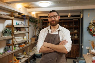 Önlüklü bir esnaf çömlek atölyesindeki kameraya bakıyor.
