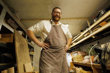 Önlüklü neşeli bir zanaatkar çömlek atölyesindeki kameraya bakıyor.