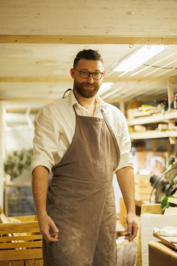 Önlüklü bir çömlekçinin portresi atölyedeki kameraya bakıyor.