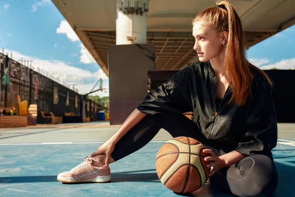 A basketball player is sitting on a sports field with a basketball and looks away. Sports, fitness, lifestyle.