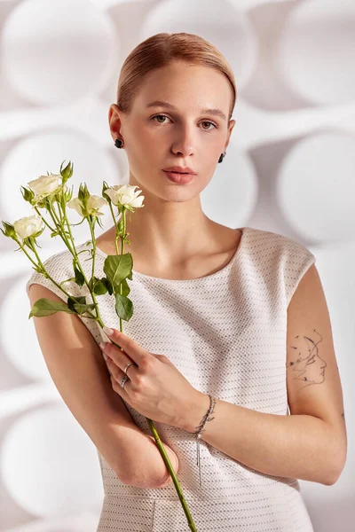 Portrait of a beautiful young disabled woman, born without an arm, holding a flower in her hands in a white dress. Diversity disability and flowers