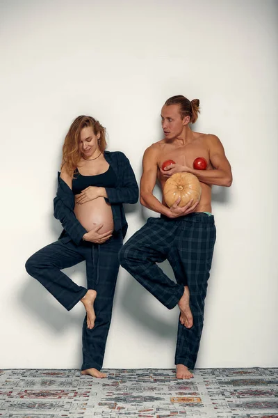 夫が遊んでいる面白い妊娠中の女の子 妊娠中の女性は彼女の腹を保持し 彼女の夫は彼の腹とトマトの代わりにカボチャを胸に持っています — ストック写真