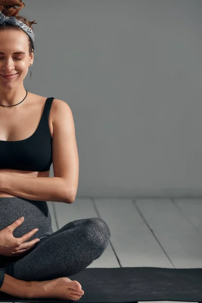 Young pregnant millennial woman sitting on the mat touches her belly after performing prenatal and meditation exercises at a yoga class. Concept of life and maternity.