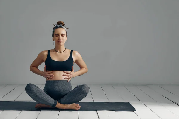 Young pregnant millennial woman sitting on the mat touches her belly after performing prenatal and meditation exercises at a yoga class. Concept of life and maternity.