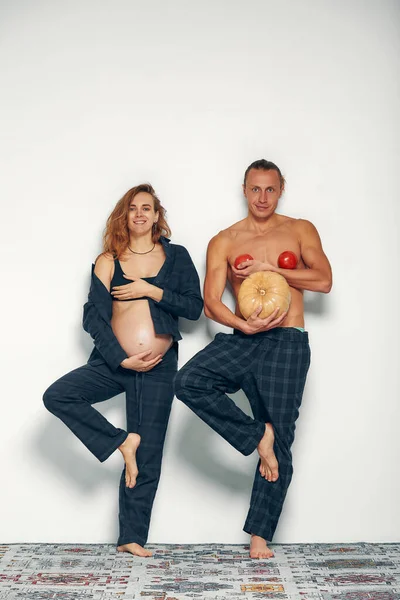 夫が遊んでいる面白い妊娠中の女の子 妊娠中の女性は彼女の腹を保持し 彼女の夫は彼の腹とトマトの代わりにカボチャを胸に持っています — ストック写真