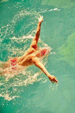 Kırmızı şapkalı bir adam havuzda yüzüyor ve güneş ışınları arasında gözlük takıyor, yüzme tekniğini, üst görüşü, kelebek stilini, kopyalama alanını eğitiyor.