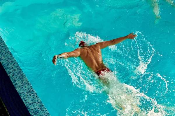 A man swims in the pool in a red cap and glasses among the rays of the sun, trains swimming technique, top view, butterfly style, copyspace