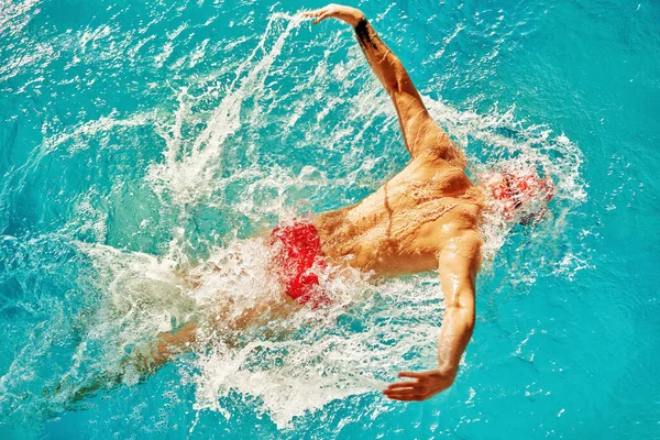 A man swims in the pool in a red cap and glasses among the rays of the sun, trains swimming technique, top view, butterfly style, copyspace