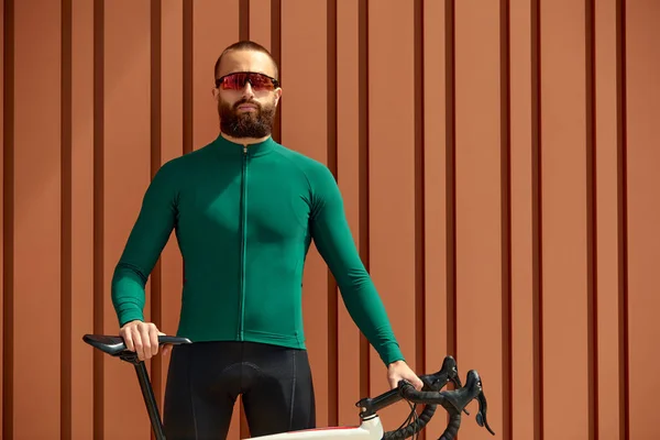 A young cyclist against the backdrop of urban architecture in bright green-black clothes against the background of a red wall with columns, training on a bicycle in the city, walking on a bicycle