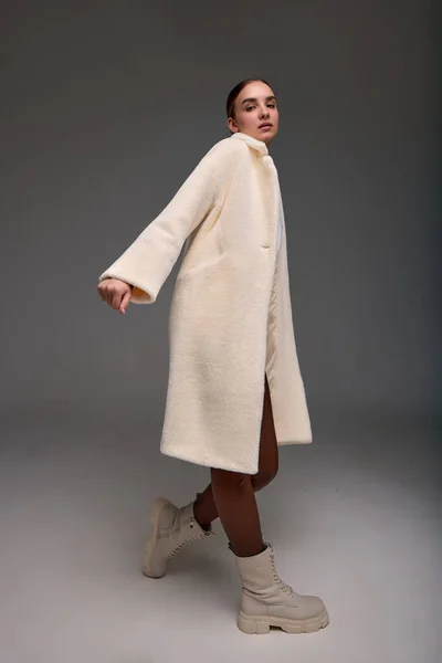 A girl in a light artificial fur coat on a gray background. Modern fashion trend for fur coats made of faux fur. Model posing in a light fur coat on a gray background.
