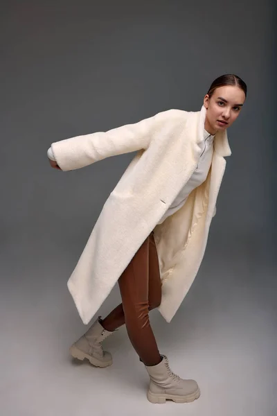 A girl in a light artificial fur coat on a gray background. Modern fashion trend for fur coats made of faux fur. Model posing in a light fur coat on a gray background.