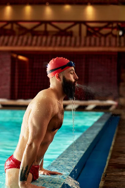 一名戴着帽子和护目镜的男游泳运动员从游泳池里跳出来 戴着红色的泳帽游泳 从游泳池里飞出 — 图库照片