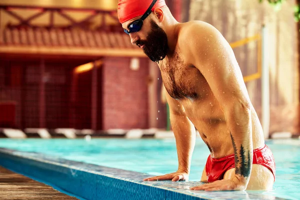 一名戴着帽子和护目镜的男游泳运动员从游泳池里跳出来 戴着红色的泳帽游泳 从游泳池里飞出 — 图库照片