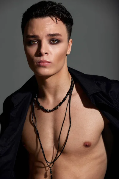 Full face studio portrait of a young teenager brunette boy in a black shirt with a naked torso and beads around his neck. Short trendy haircut. Elegant sexy style. Gray background