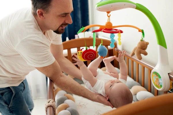 一个快乐的爸爸把他的孩子和一个音乐玩具放在一个婴儿床里 父亲抱着他的孩子 快乐的童年 — 图库照片