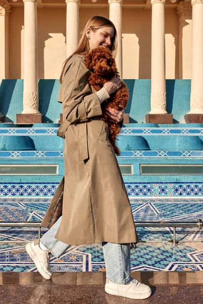一个身穿米色风衣和牛仔裤的年轻金发女子的全长肖像 她走路时怀里抱着一只红色的玩具狮子狗 在街上带着宠物散步 — 图库照片