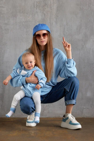 一个穿着牛仔裤 蓝色毛衣 滑稽的蓝色帽子和太阳镜的年轻时髦妈妈坐在她的发髻上 抱着她微笑的孩子 宝宝和妈妈穿一样的颜色衣服 工作室拍摄 灰色背景 — 图库照片