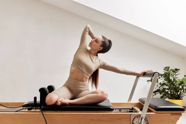 A young girl is doing Pilates on a reformer bed in a bright studio. A beautiful slender brunette in a beige bodysuit does exercises to strengthen the spine and back muscles