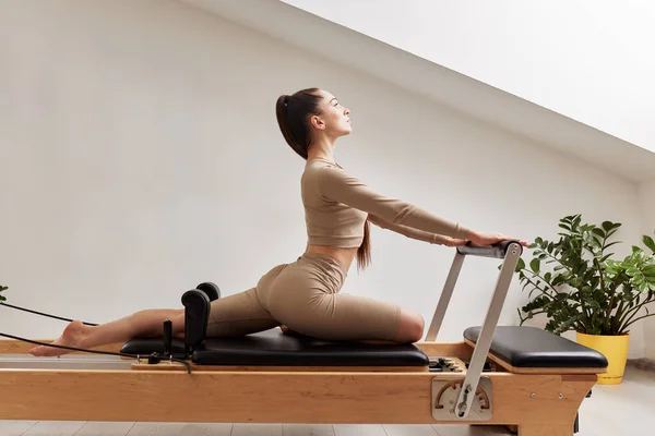 A young girl is doing Pilates on a reformer bed in a bright studio. A beautiful slender brunette in a beige bodysuit does exercises to strengthen the spine and back muscles