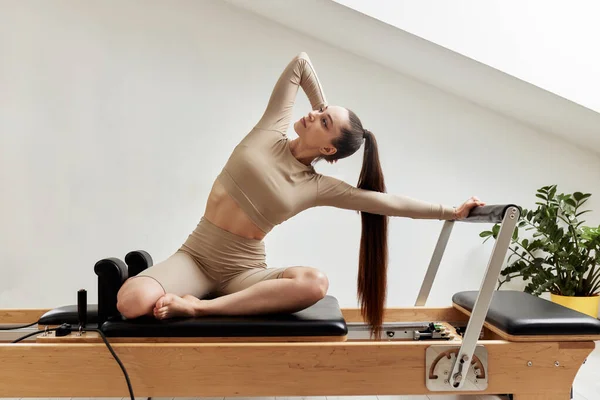 A young girl is doing Pilates on a reformer bed in a bright studio. A slender brunette in a beige bodysuit makes bends to strengthen the spine and back muscles. Healthy lifestyle concept