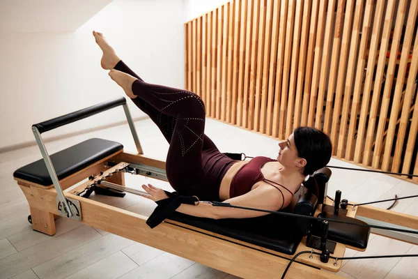 一个年轻的女孩正在一间明亮的工作室里的一个改革者床上做普拉提 一个纤细的黑发女人在仰卧时做运动来锻炼肌肉 健康生活方式概念 — 图库照片