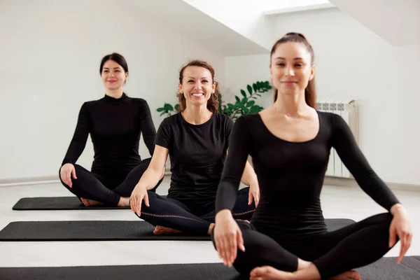 若い女性が明るいスタジオでピラティスをやっています 黒のスーツを着た3人のスレンダーな笑顔のスポーツウーマンはヨガマットに座って練習をします 健康的なライフスタイルのコンセプト — ストック写真