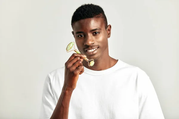 African guy in a white t-shirt with jade de-puffing face roller. Black man in studio with beauty, health and natural skin procedure. Wellness, cosmetics and skin care for men. Close up portrait