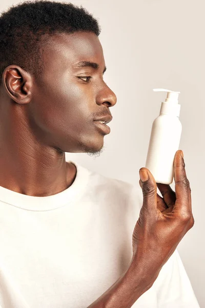 African guy in a white t-shirt with a bottle of lotion or skin cream. Black man in studio with beauty, health and natural skin procedure. Wellness, cosmetics and skincare for men. Close up portrait