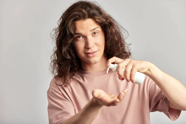 Young handsome smiling guy with long brown hair squeezing skincare cream from the dispenser to his hand. Caucasian man in studio with healthy skin procedure. Wellness, cosmetics and skincare for men