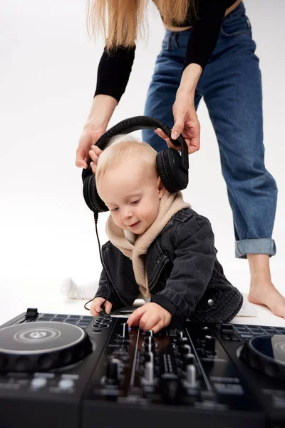 stock image A cute kid in a beige hoodie and a funny black motorcycle jacket sits on the floor with a dj mixing console. A barefoot mom in blue jeans puts DJ headphones on his head.