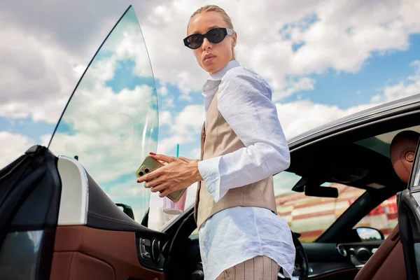 一位身穿白衬衫和米色背心的年轻漂亮的女商人站在她的豪华轿车旁边 在商务中心或购物中心的停车场 一个戴着太阳镜的自信的金发女人 — 图库照片