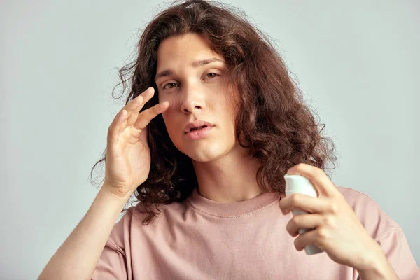 A handsome guy with long brown hair doing self-care routine using a facial moisturizer in a studio. Caucasian man caring for the skin to maintain his youthful and healthy appearance. Mens skincare