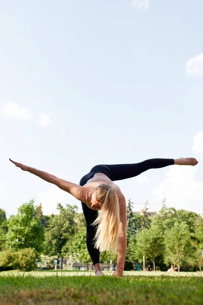Young woman practice yoga in summer park. Slender caucasian blonde girl in black bodysuit doing exercises to strengthen her body and spirit. Morning gymnastics in fresh air. Healthy lifestyle
